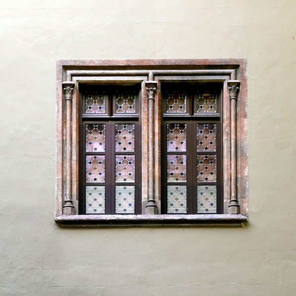 La Seu d’Urgell.- Palau Episcopal refet en el S. XIX – Finestra del pati emmarcada per columnes amb capitells decorats amb caps i la del centre només amb decoració vegetal (9/2026)